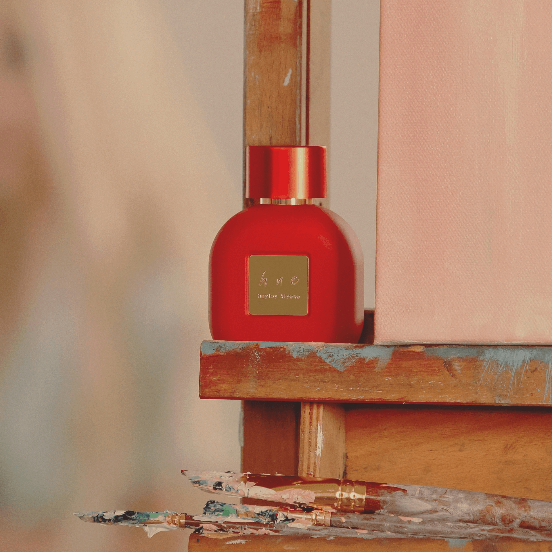 Hue by Hayley perfume created by female celebrity, Hayley Kiyoko. The fragrance is perched on wooden stand with paint brushes strategically placed around it.